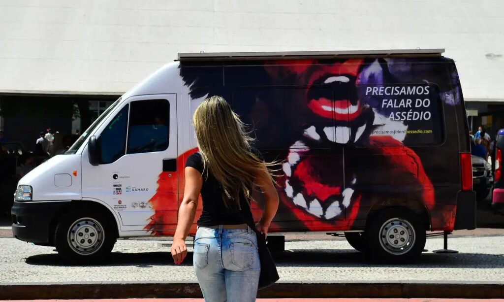 Mulher de costas em frente a van com arte de boca aberta e campanha "Precisamos Falar".