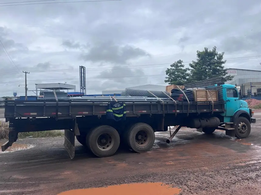 Caminhão de carga com tubos metálicos, telas e caixas em pátio industrial.