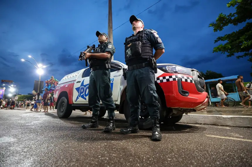 Dois policiais militares uniformizados com coletes e um fuzil, ao lado de uma viatura em patrulha noturna.