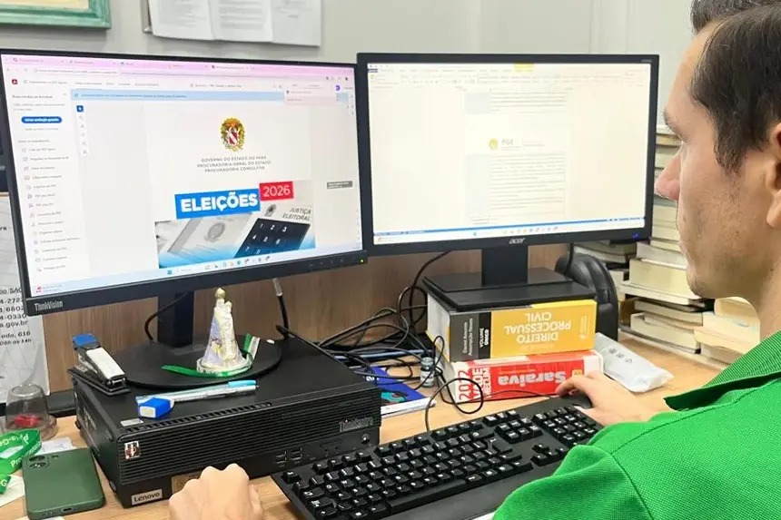 Pessoa trabalhando em computador com dois monitores, exibindo site sobre eleições 2026 e documento legal, com livros de Direito.