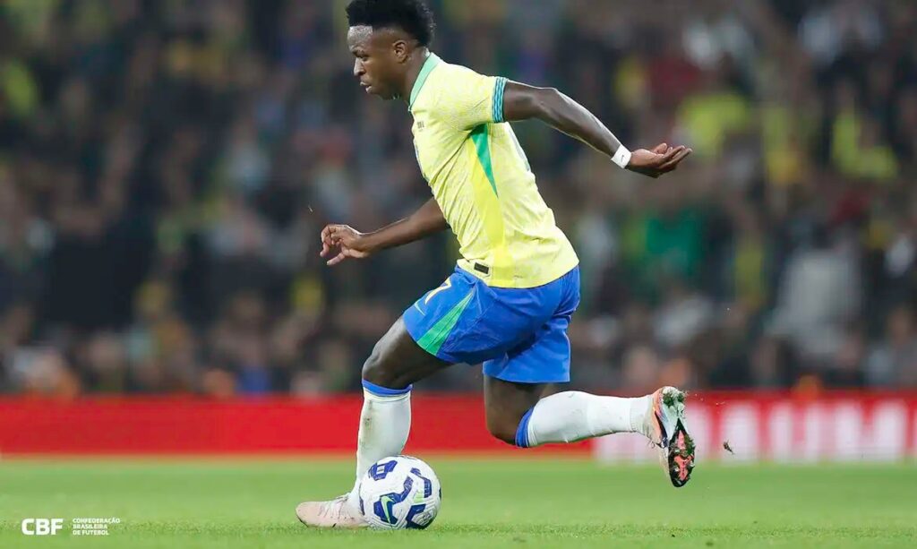 Vinicius Junior em campo com a camisa da seleção brasileira, driblando uma bola.
