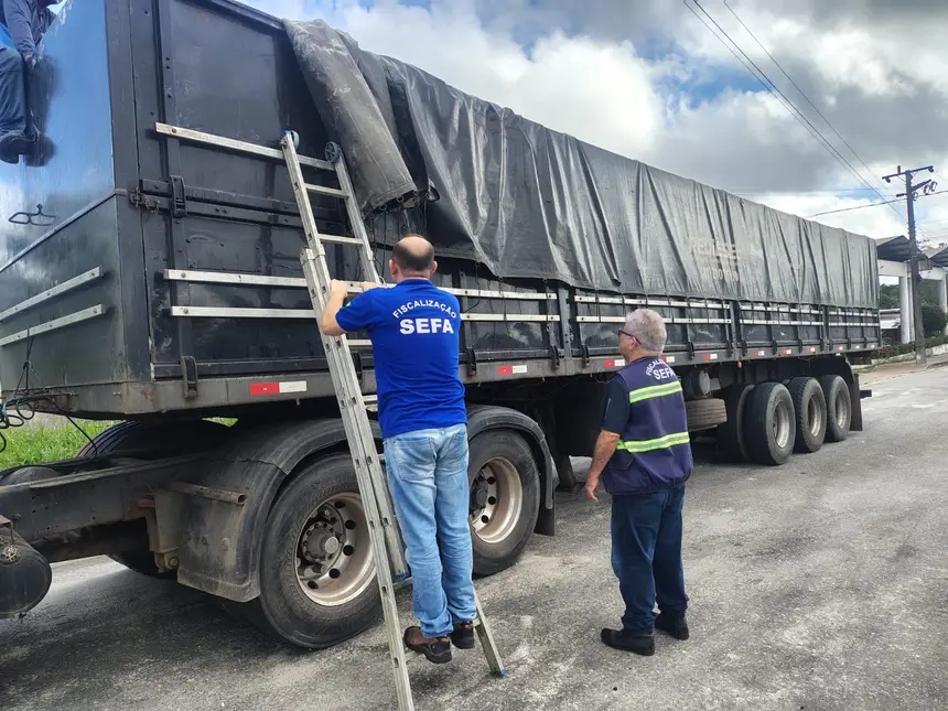 Dois agentes da SEFA fiscalizando a carga de um caminhão com lona preta.
