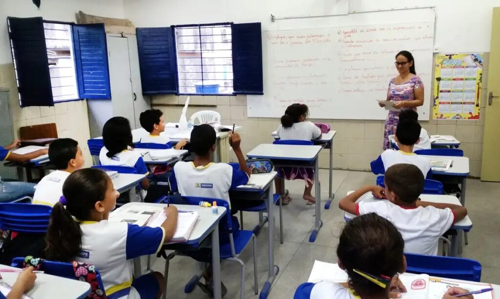Uma sala de aula com diversos alunos em uniformes azuis e brancos, sentados em carteiras, e uma professora de pé à frente. Um aluno levanta a mão para participar.