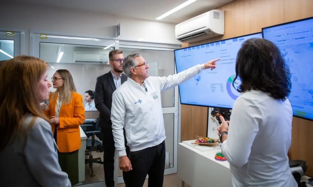 Homem de jaleco branco apontando para tela com gráficos e dados em sala de reunião.