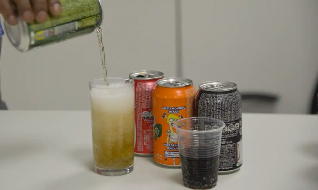 Bebida sendo servida em copo ao lado de latas de refrigerante variadas.