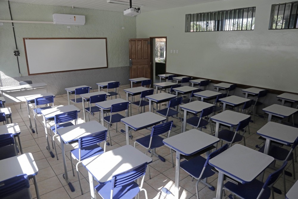 Sala de aula vazia com carteiras azuis e brancas, lousa, ar condicionado e projetor.