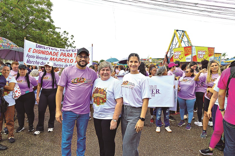 Homem e duas mulheres sorrindo em primeiro plano, com um grupo de pessoas e uma faixa 'Direitos, Respeito e Dignidade' ao fundo, durante uma marcha.