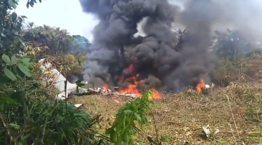 Destroços de avião em chamas e fumaça em área de mata.