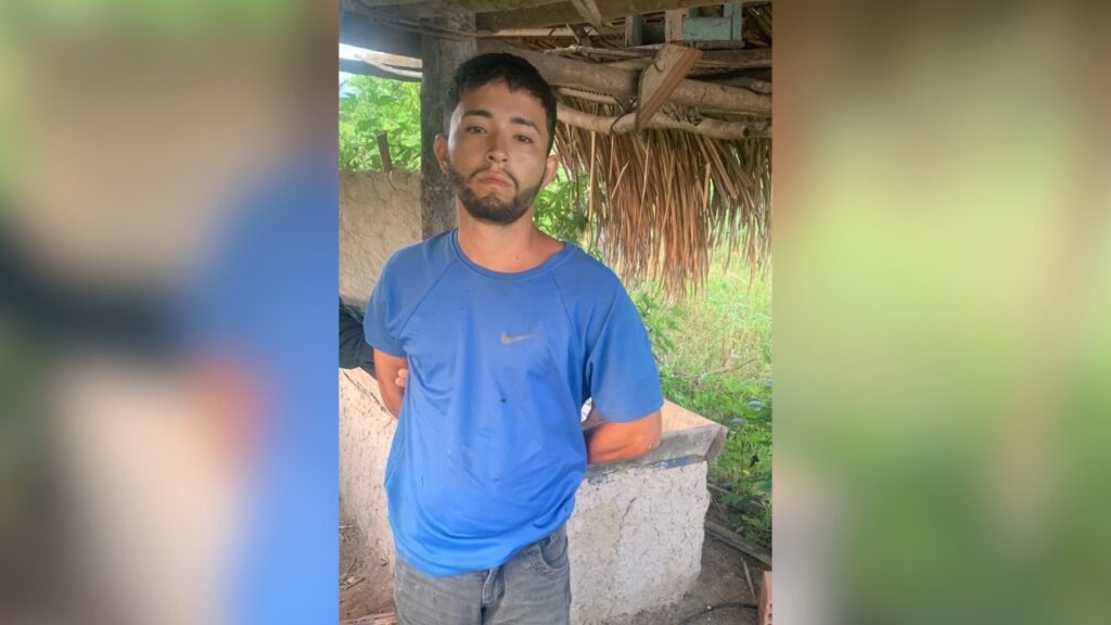 Jovem com barba e camiseta azul em ambiente rústico.