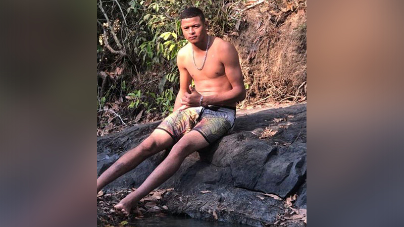 Jovem sem camisa e de bermuda florida, sentado em uma rocha à beira de um riacho, fazendo um sinal de positivo.