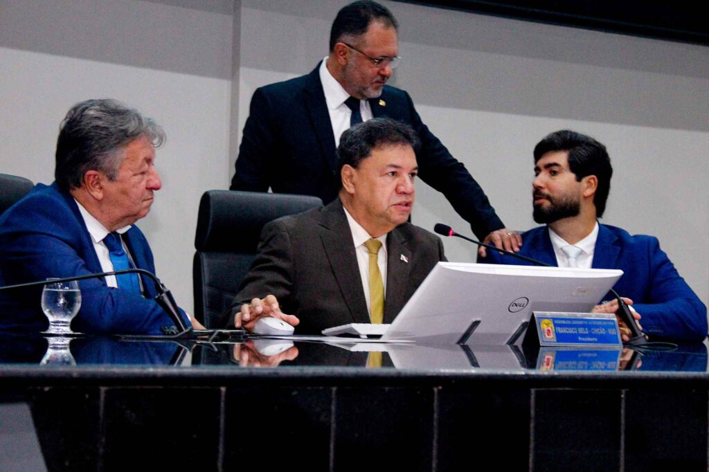 Grupo de quatro homens em uma reunião formal. O orador central usa terno e gravata dourada.