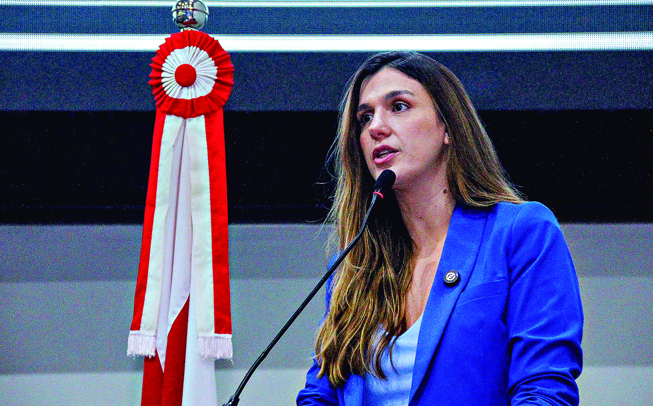 Mulher de blazer azul falando ao microfone em evento.