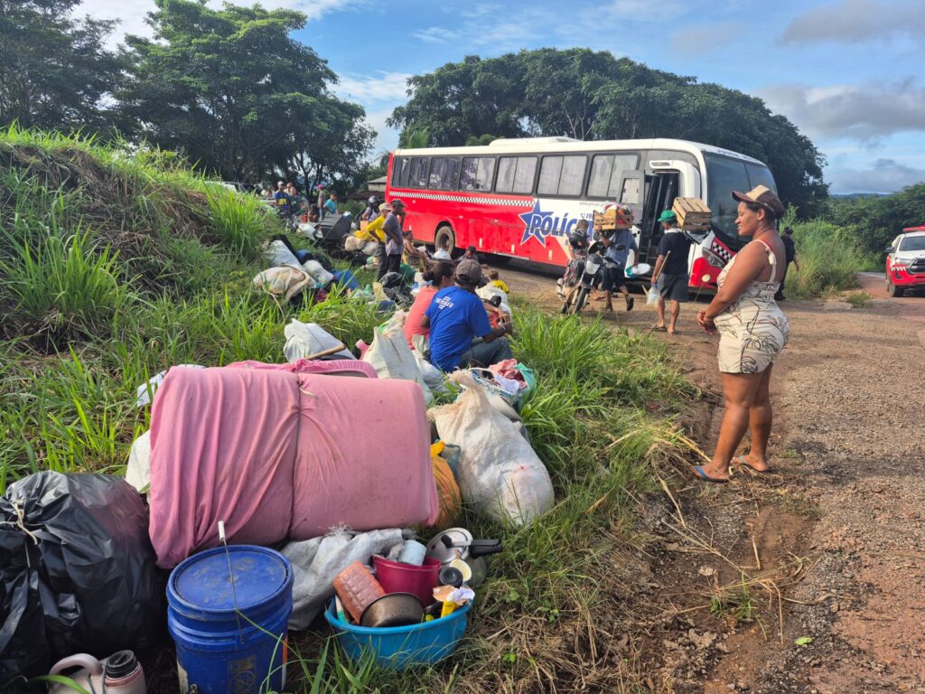 Ônibus vermelho com 'Polícia' parado em estrada de terra, pessoas e pertences espalhados na grama.