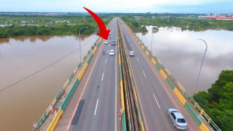 Vista aérea de uma ponte movimentada sobre um rio marrom com carros e trabalhadores.