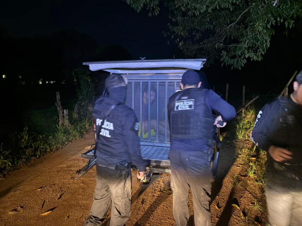 Dois policiais civis próximos a um veículo de transporte com um detido em compartimento gradeado, à noite.