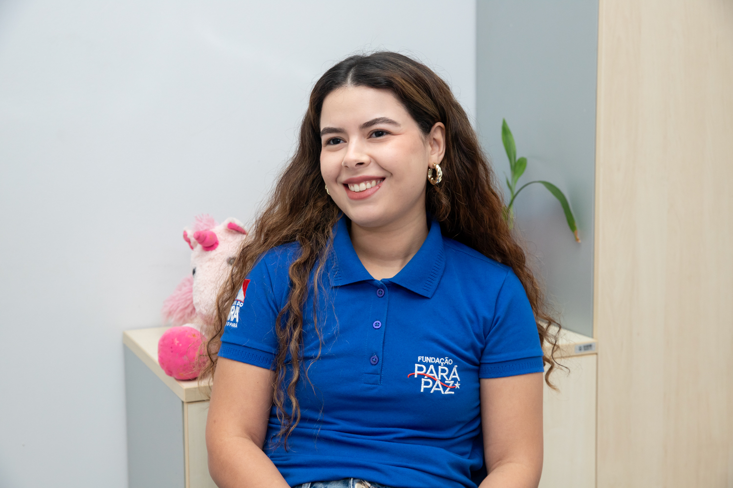 Jovem mulher sorrindo em camisa polo azul da Fundação Para Paz, com cabelo ondulado e brinco dourado.