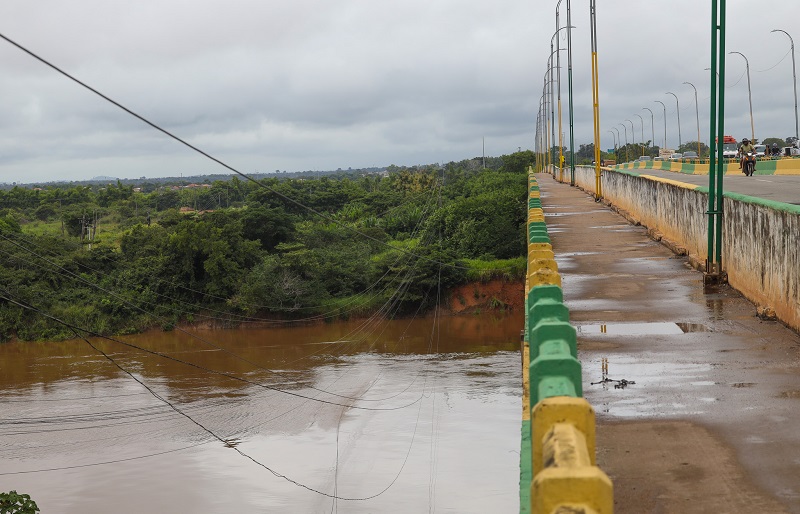 Ponte sobre rio barrento com mata densa e fiação exposta, sob céu nublado.