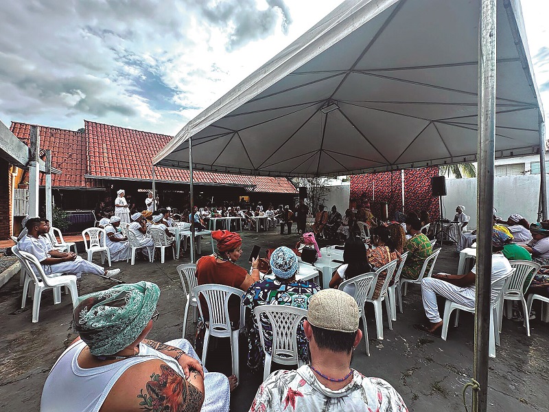 Pessoas sentadas em encontro cultural ao ar livre, muitas vestindo branco e turbantes sob uma tenda.