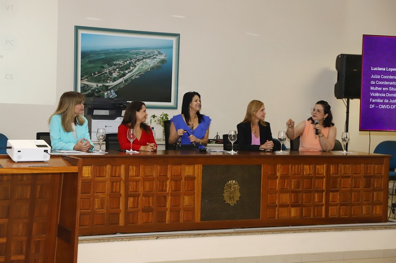 Cinco mulheres em um painel de discussão, com uma juíza falando ao microfone e um telão exibindo seu nome e cargo.