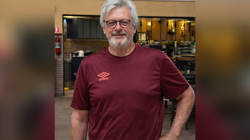 Homem de cabelos e barba grisalhos sorrindo, usando óculos e camiseta Umbro vinho.