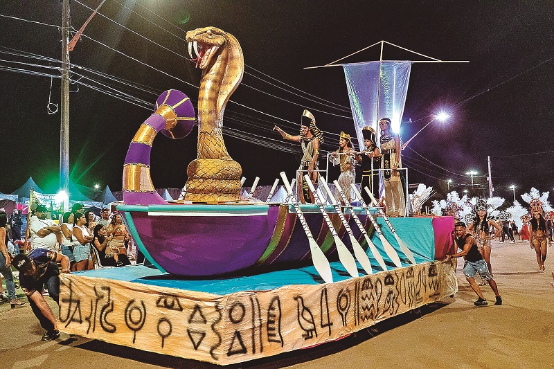 Carro alegórico egípcio com cobra dourada gigante e pessoas fantasiadas em desfile noturno.