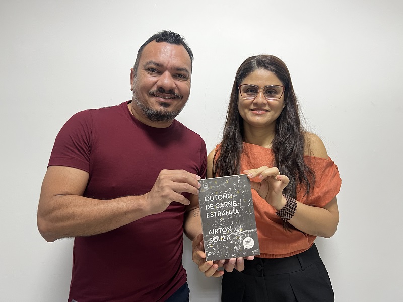 Homem e mulher sorrindo segurando o livro "Outono de Carne Estranha" de Airton Souza.