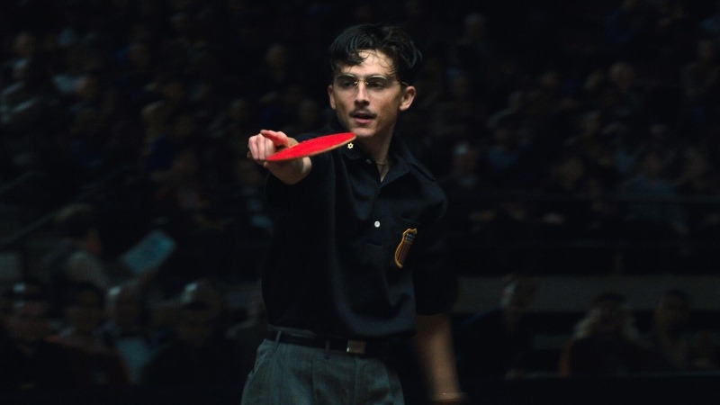 Homem jovem com óculos e bigode, vestindo polo escuro, segurando uma raquete de ping-pong vermelha e apontando-a para frente.