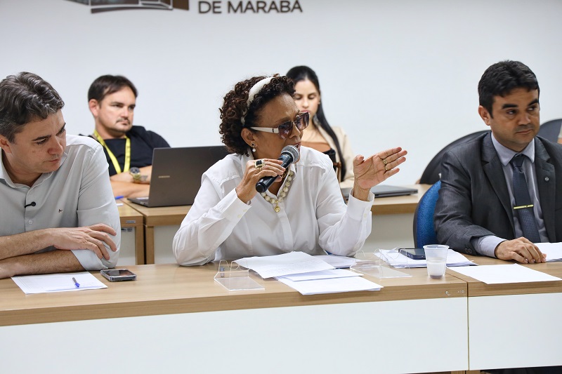 Mulher com óculos escuros e microfone em reunião, gesticulando.