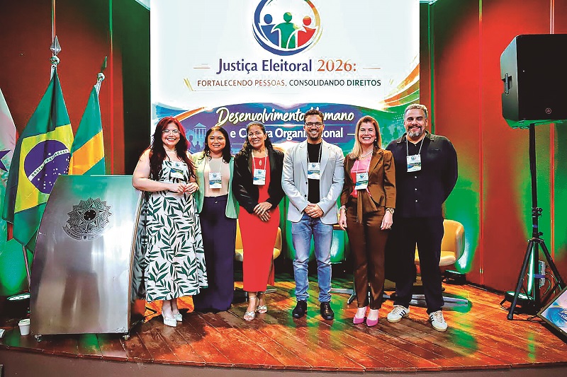 Seis profissionais posam em palco com bandeiras do Brasil e painel institucional da Justiça Eleitoral 2026.