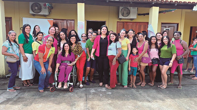 Grupo de mulheres de diversas idades, incluindo uma em cadeira de rodas, sorrindo em frente a um prédio amarelo.