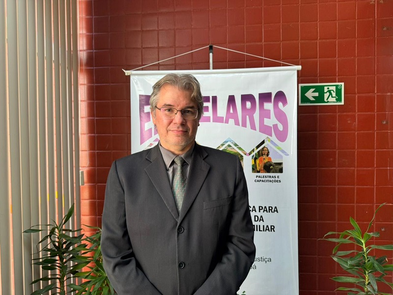 Homem de óculos e terno escuro em frente a um banner de evento.