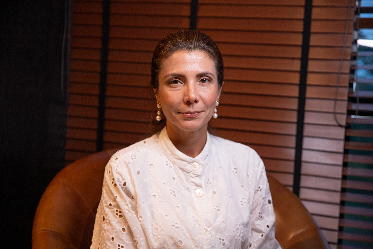 Retrato de mulher de meia-idade com blusa branca bordada e brincos de pérola, sentada em poltrona de couro.