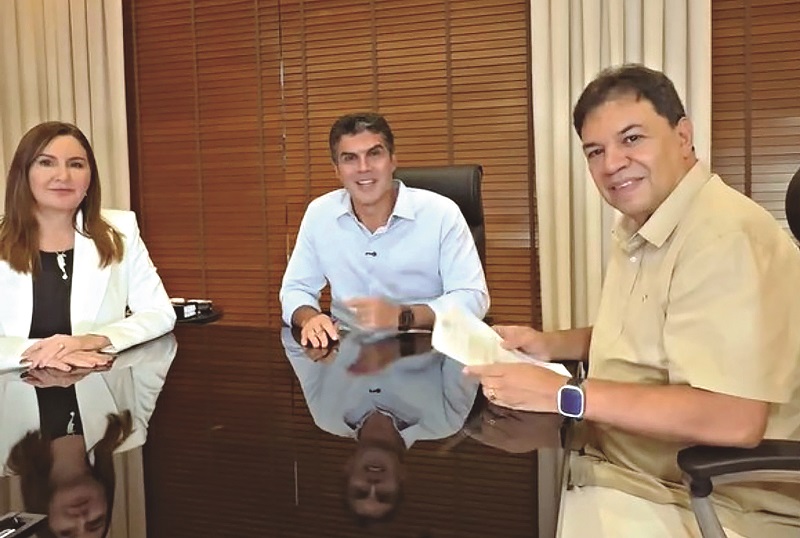 Três pessoas, uma mulher e dois homens, sorrindo em uma mesa de reunião com persianas de madeira ao fundo.
