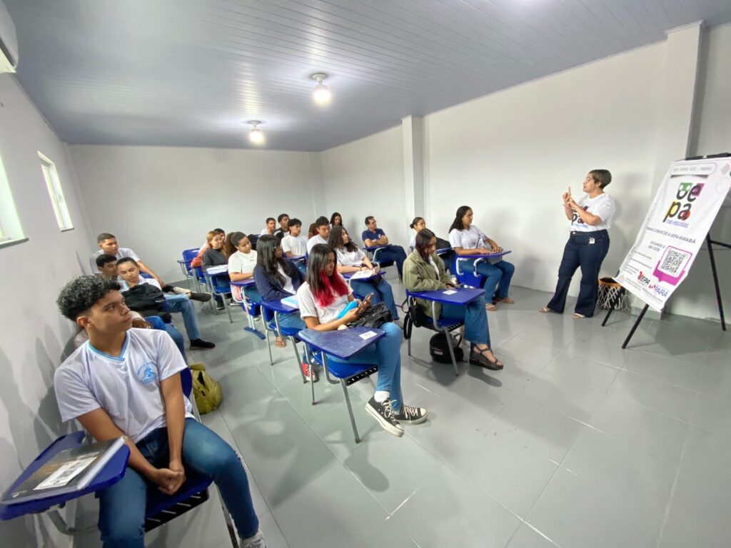 Mulher apresentando para estudantes em sala de aula com carteiras azuis.