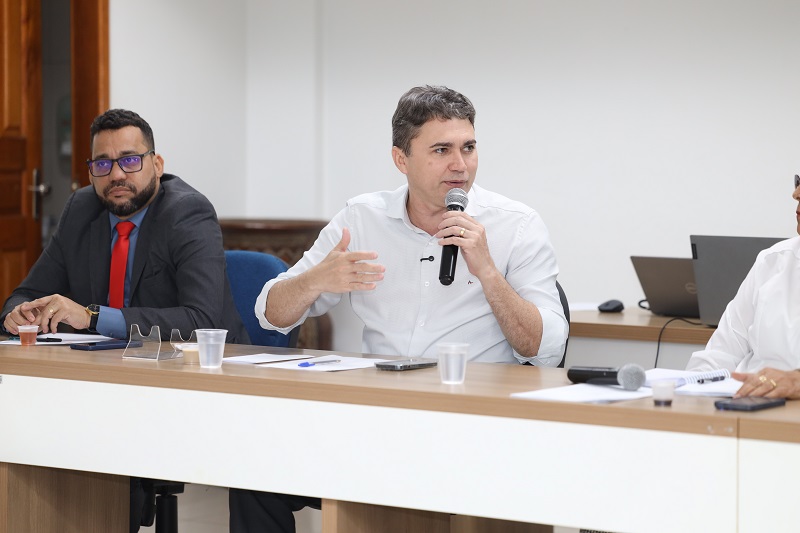 Homem falando ao microfone em uma reunião com outros participantes sentados à mesa.