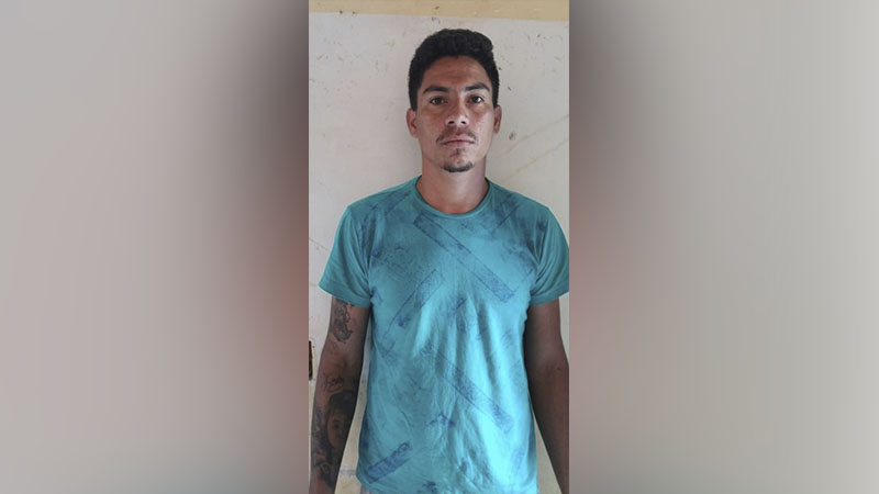 Homem de camiseta azul-petróleo com tatuagens nos braços, em pé contra uma parede clara.