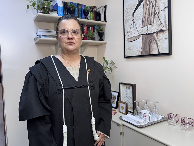 Mulher de toga e óculos em escritório com estante de livros e quadro da balança da justiça.