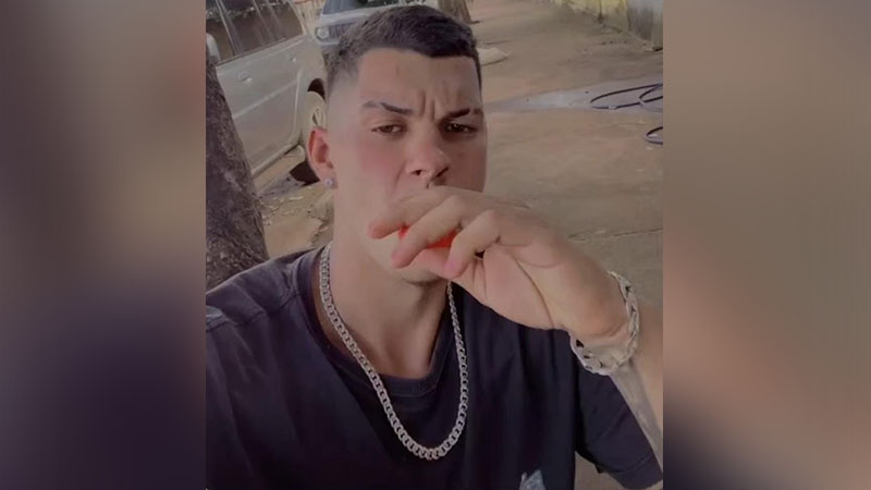 Homem jovem com cabelo curto e corrente prateada, com a mão na boca, em ambiente externo.