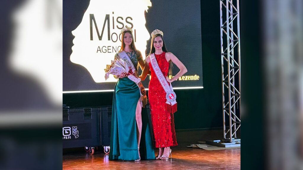 Duas mulheres, uma de vestido verde e outra de vermelho, com coroas e faixas de miss em um palco de evento.