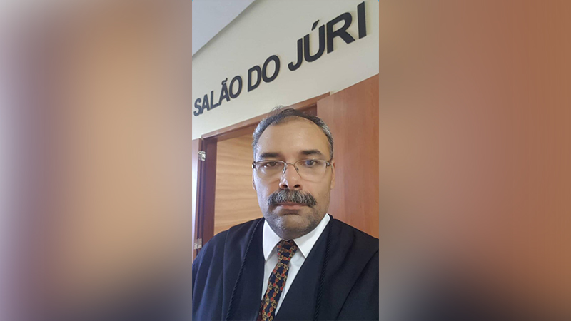 Homem de toga com óculos e bigode em frente a uma porta com a placa 'SALÃO DO JÚRI'.