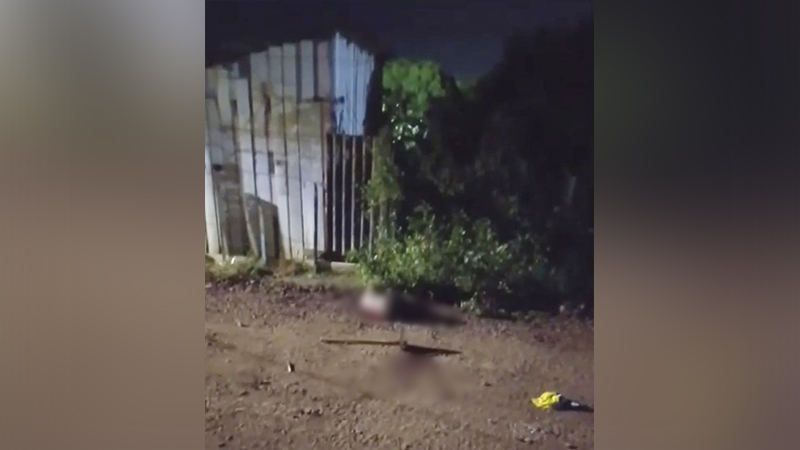 Cena noturna com partes de um corpo e uma arma no chão, em área rural ou degradada.