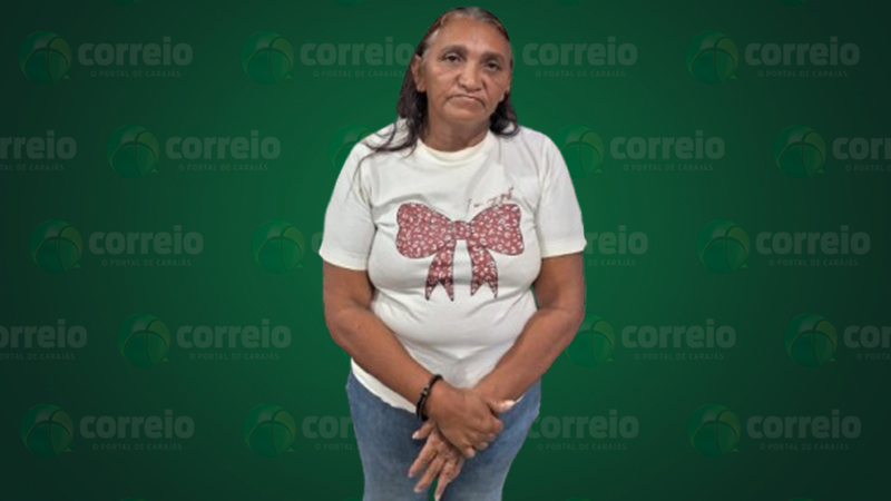 Uma mulher de meia-idade com cabelos escuros, camiseta branca estampada com laço vermelho e calça jeans, posando para a câmera.