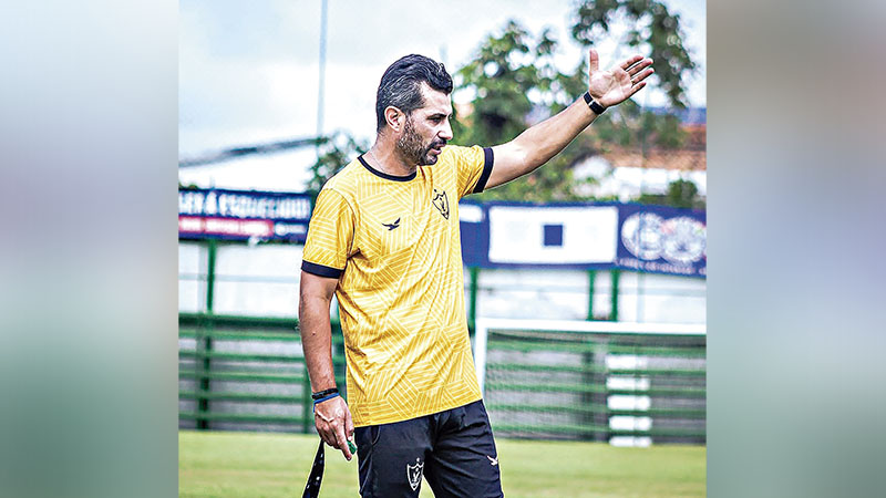 Treinador de futebol em campo, vestindo uniforme amarelo, gesticulando.