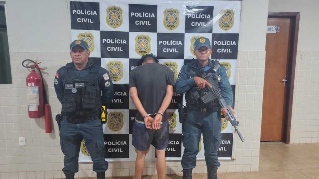 Homem algemado de costas entre dois policiais civis fardados em frente a um banner da Polícia Civil.