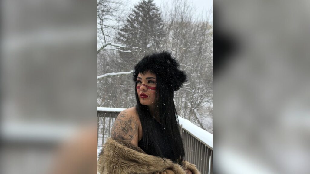 Mulher com maquiagem tribal, chapéu de pele e casaco, em meio à neve caindo.