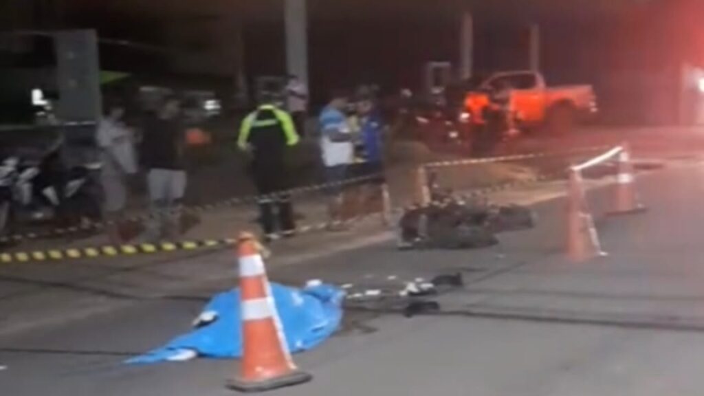 Cena noturna de um incidente grave com corpo coberto, cone de trânsito e pessoas no local.