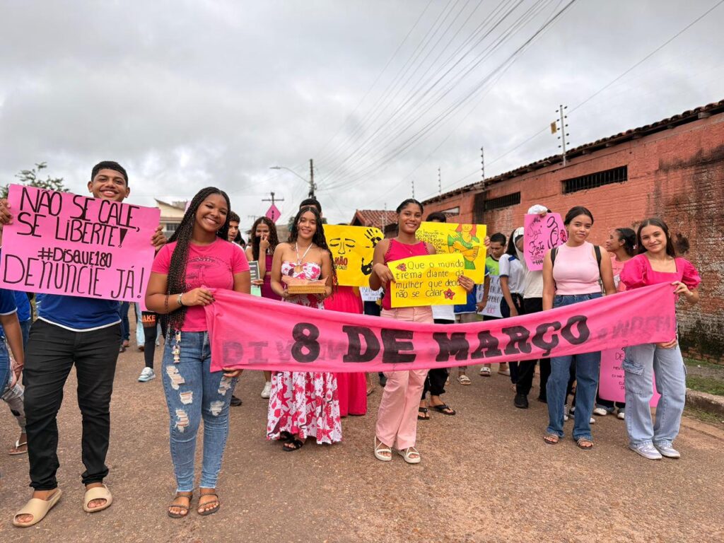 Grupo de jovens, principalmente mulheres, em uma passeata segurando cartazes e uma faixa rosa escrito "DIA 8 DE MARÇO" para o Dia Internacional da Mulher.