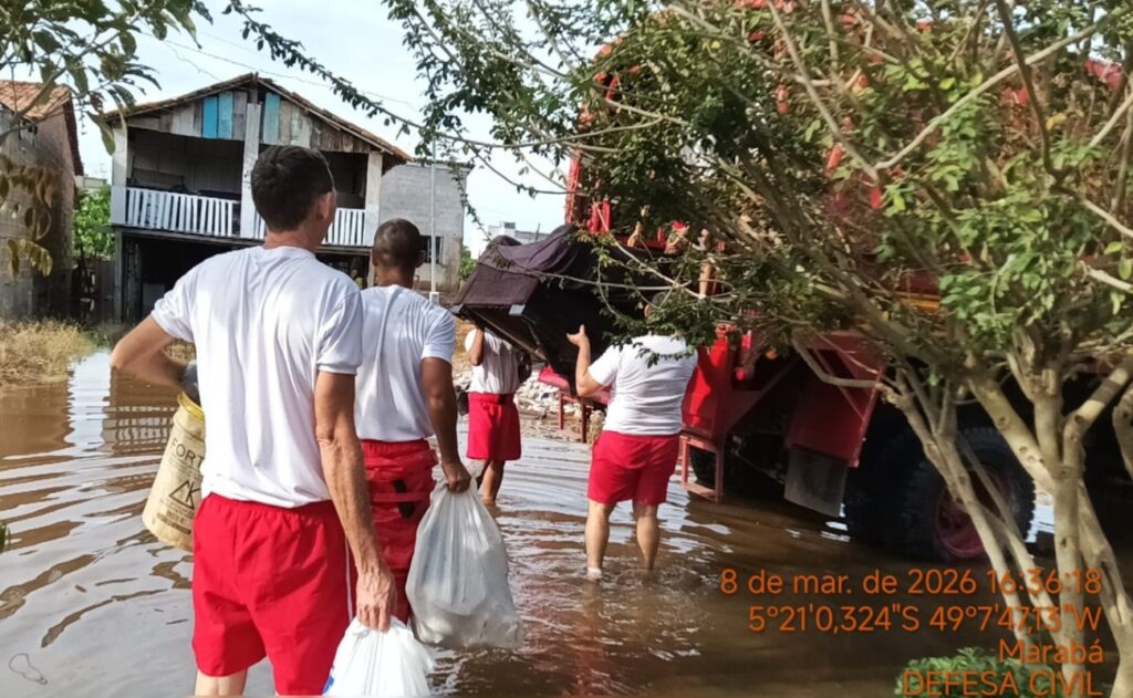 Homens em roupas brancas e vermelhas resgatando pertences em área alagada, com um caminhão tombado ao fundo.