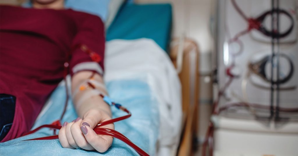 Pessoa em cama hospitalar recebendo tratamento de hemodiálise com tubos de sangue visíveis.