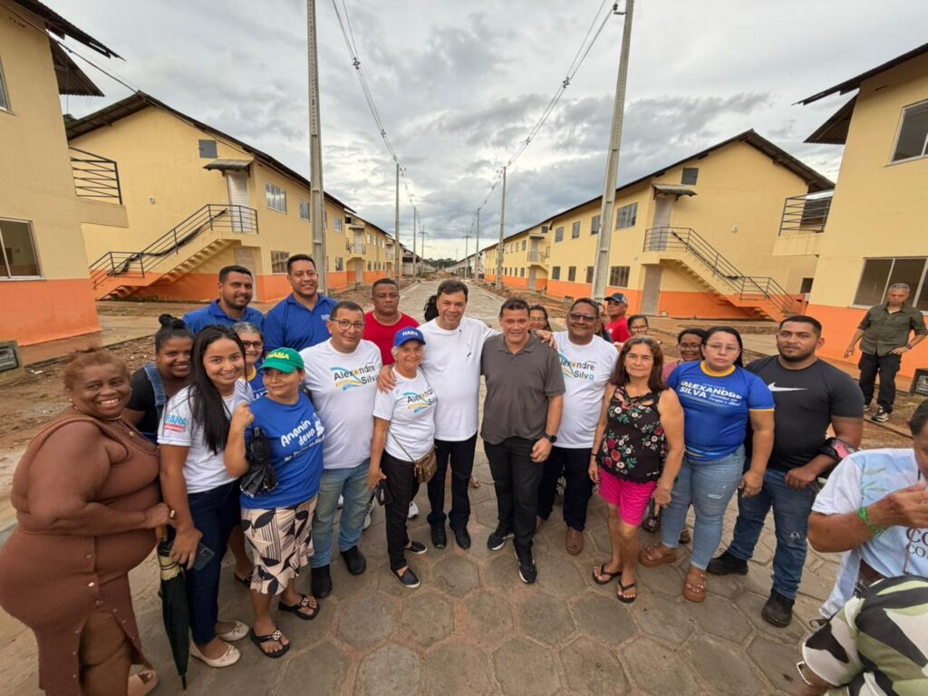Grupo de pessoas, incluindo o político Alexandre Silva, posando em frente a casas populares de cor amarela em uma rua no Brasil.